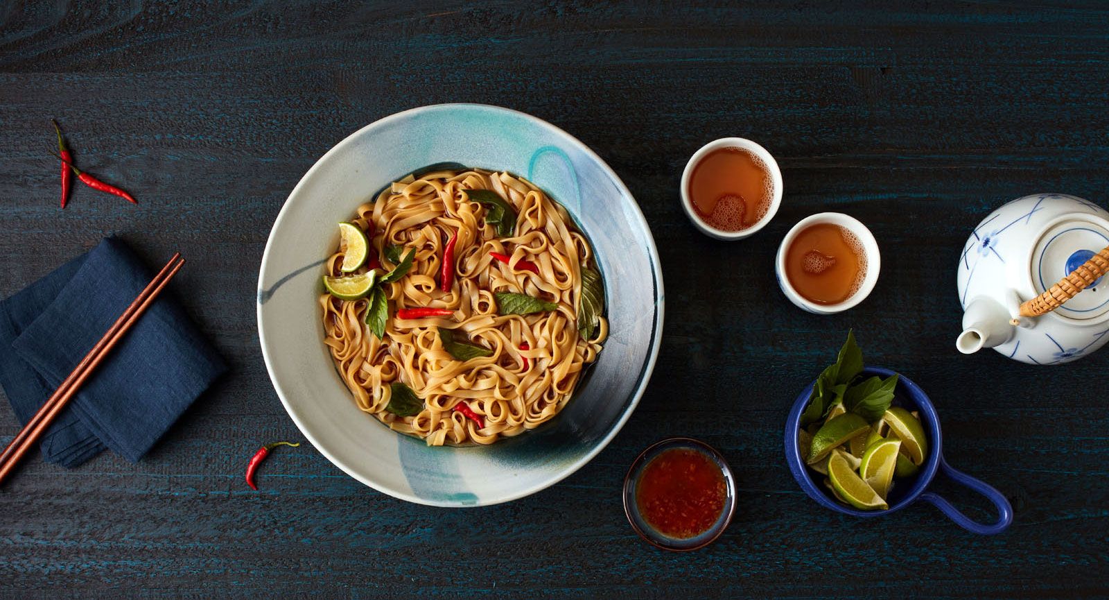 a bowl of flat egg noodles garnished with lime wedges and whole spicy peppers on a table with chopsticks, a teapot and bowls of various sauces