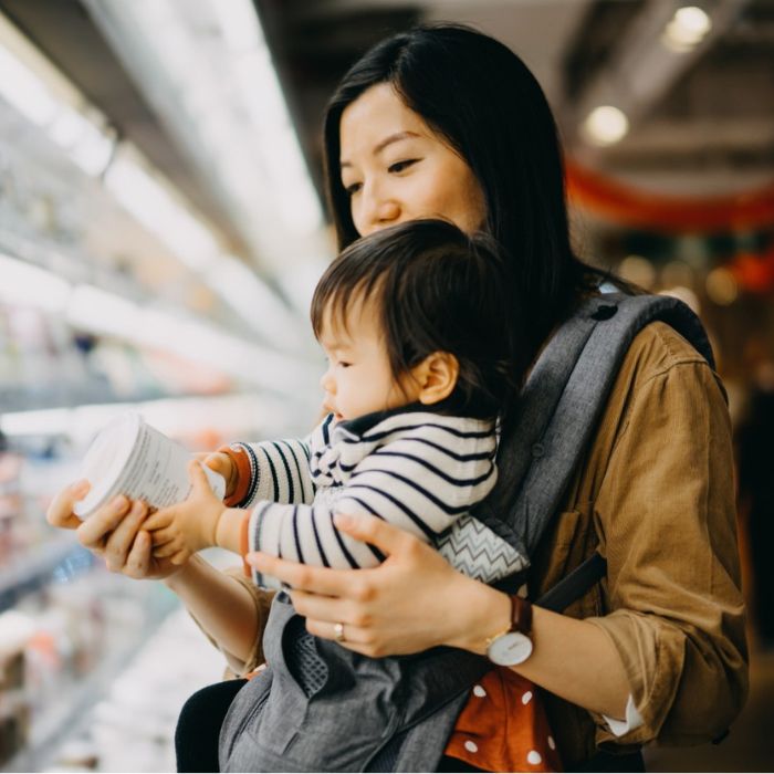 woman reading label on food container in grocery store aisle while holding infant