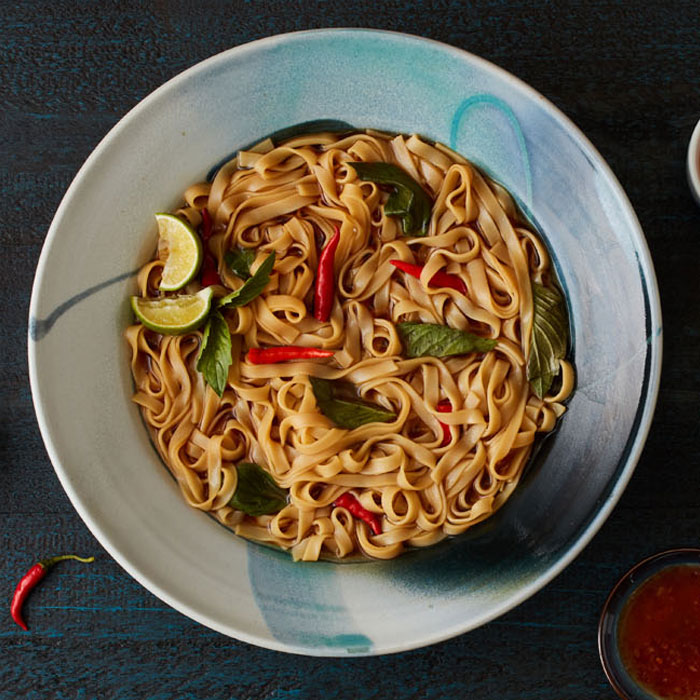 A bowl of flat egg noodles that contains several small peppers and two lime wedges